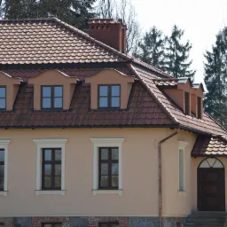 Elewacja domu z beżową fasadą, brązowymi oknami i czerwoną dachówką. Widoczne detale architektoniczne, takie jak lukarny i kamienne wykończenie.