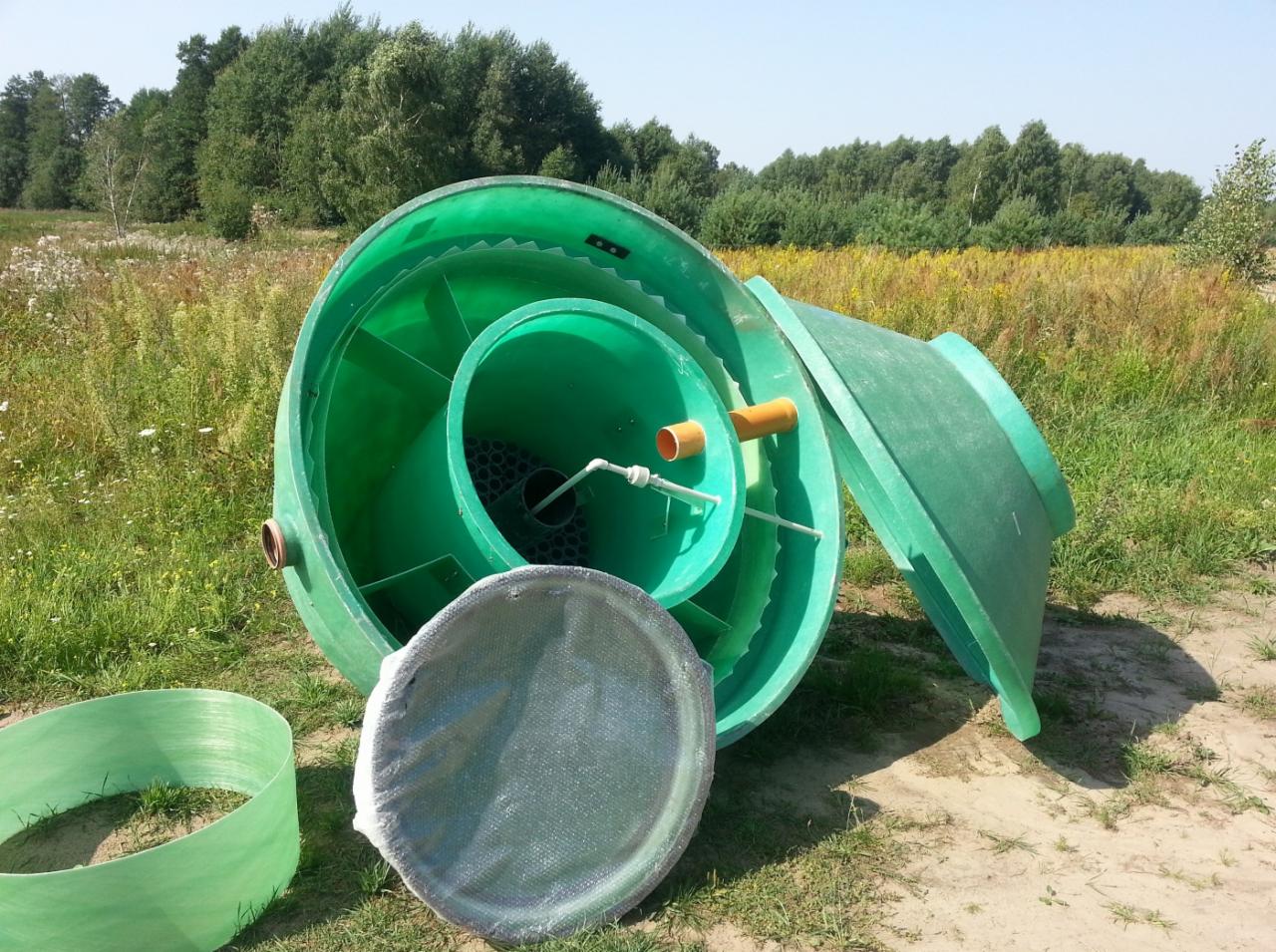 Elementy składowe zielonej, plastikowej oczyszczalni ścieków rozłożone na trawie przed montażem, widoczny zbiornik z rurami, pokrywa i filtr.