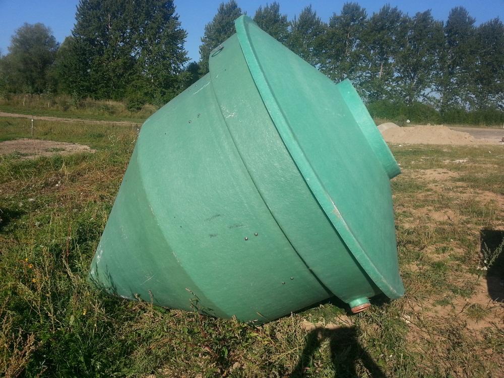 Zielony, stożkowy zbiornik z tworzywa sztucznego leży na trawie w słoneczny dzień, widoczny cień osoby robiącej zdjęcie, w tle drzewa i usypana ziemia.