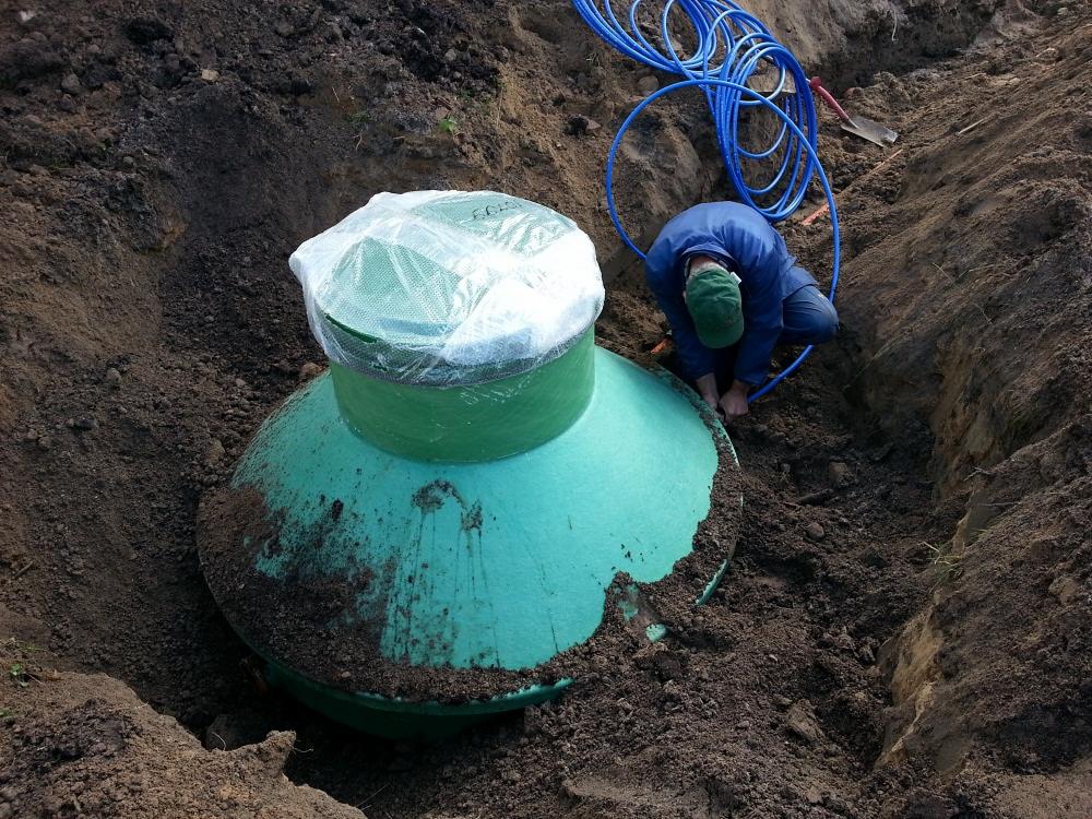 Montaż zielonego zbiornika z tworzywa sztucznego w wykopie ziemnym; widoczne niebieskie rury i pracownik w niebieskim kombinezonie podłączający instalację.