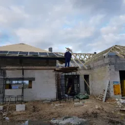 Budowa domu z bloczków: widoczne ściany w trakcie murowania, częściowo wykonane drewniane elementy dachu, robotnik na rusztowaniu z drewnianymi belkami w rękach, materiały budowlane składowane obok...