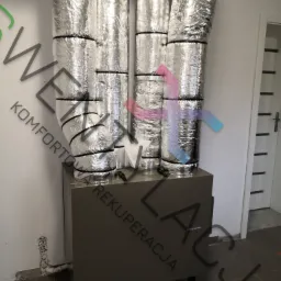 Instalacja rekuperacji z czterema rurami izolowanymi folią aluminiową, podłączonymi do szarej skrzynki rekuperatora w pomieszczeniu z widoczną instalacją hydrauliczną przy ścianie i białymi...