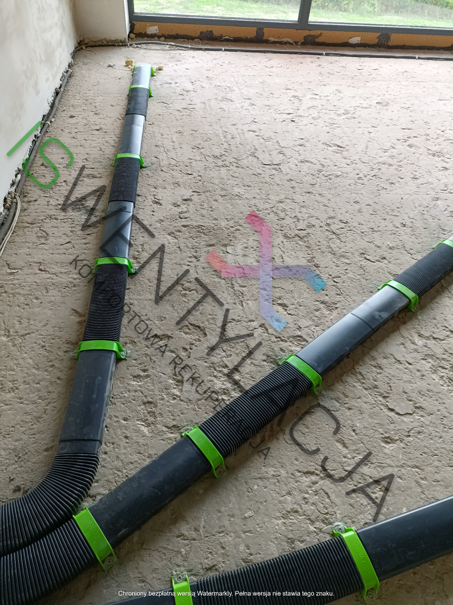 Fragment podłogi w trakcie montażu instalacji rekuperacji z widocznymi przewodami wentylacyjnymi z czarnego tworzywa, połączonymi zielonymi uchwytami, na surowej, betonowej posadzce w jasnym...