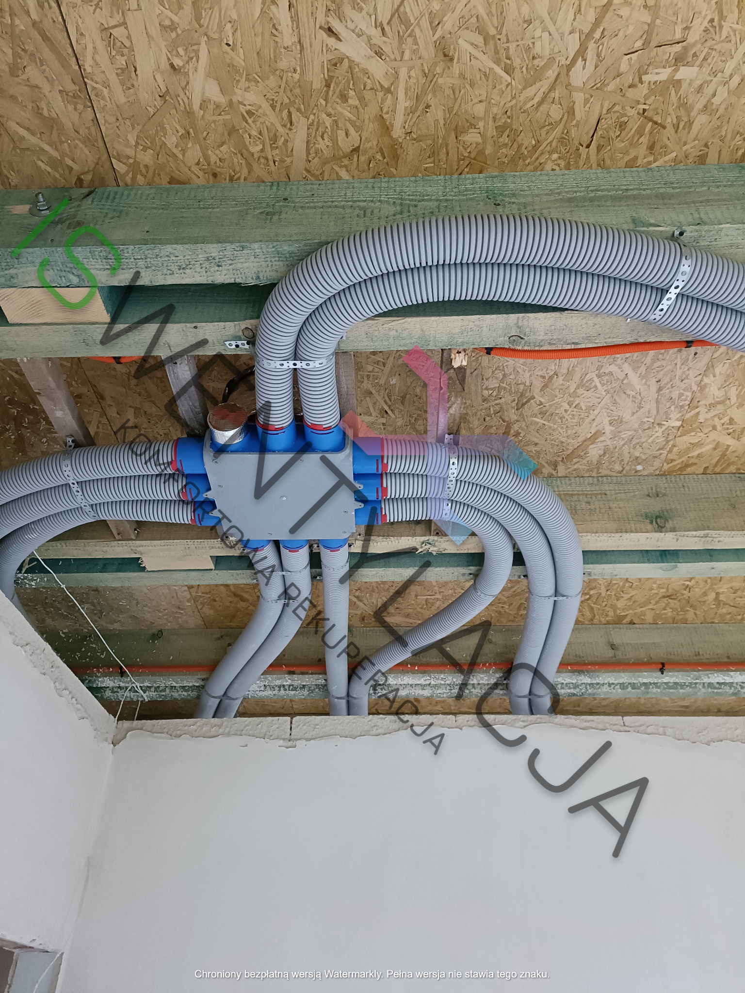 Rozprowadzenie szarych, karbowanych rur wentylacyjnych z centralnej skrzynki rozdzielczej pod drewnianym stropem, widoczne pomarańczowe peszle z przewodami elektrycznymi.