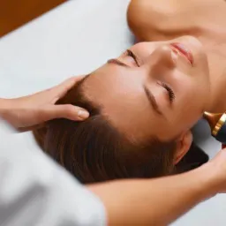 Kobieta leżąca na plecach podczas zabiegu na twarz z użyciem urządzenia z metalową końcówką, dłoń terapeuty dotyka jej skroni.