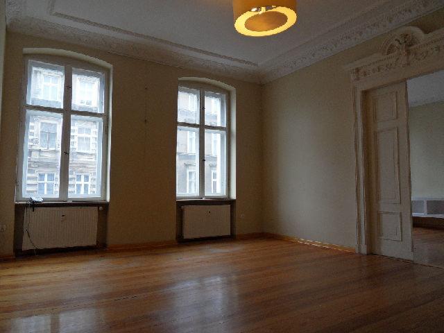 Puste wnętrze z drewnianą podłogą, dwoma dużymi oknami z widokiem na budynki i grzejnikami pod nimi, oraz ozdobnym portalem drzwiowym prowadzącym do kolejnego pomieszczenia, oświetlone wiszącą lampą.