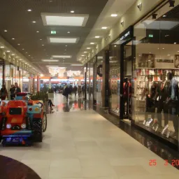 Polerowana, jasna posadzka w galerii handlowej, odbijająca światło sufitowych lamp; w tle sklepy odzieżowe i spacerujący ludzie.