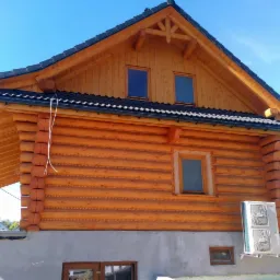 Drewniany dom z bali z zamontowanym klimatyzatorem na betonowym fundamencie, czarne dachówki, błękitne niebo w tle.