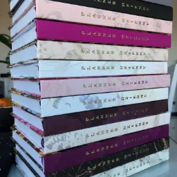 Stos kilkunastu różnorodnych, zamkniętych planerów dziennych z twardą oprawą, ułożonych jeden na drugim, z widocznymi złotymi napisami na grzbietach.