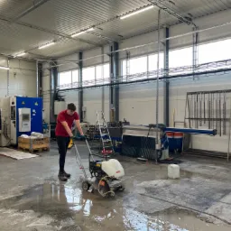 Pracownik w czerwonej koszulce obsługuje przecinarkę do betonu na mokrej posadzce w hali produkcyjnej, widoczne maszyny i narzędzia.