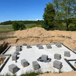 Przygotowany fundament z izolacją przeciwwilgociową i ułożonymi bloczkami betonowymi na paletach, teren wyrównany, w tle zielone pola i las pod błękitnym niebem.