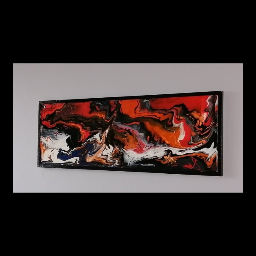 Abstrakcyjny obraz w czarnej ramie, z dominującymi kolorami czerwonym, pomarańczowym, czarnym i białym, z akcentami granatu, wykonany techniką fluid painting, zawieszony na szarej ścianie.