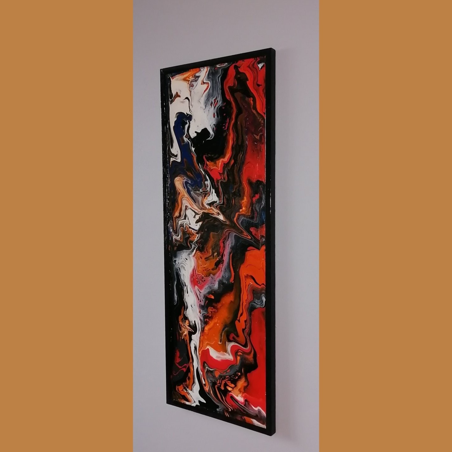 Wertykalny obraz w czarnej ramie z abstrakcyjną kompozycją farb w odcieniach czerwieni, bieli, czerni i granatu, wiszący na szarej ścianie na tle pomarańczowej ściany.