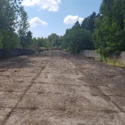 Ujęcie z perspektywy drogi zbudowanej z betonowych płyt, otoczonej z obu stron betonowym murem i drzewami, pod błękitnym niebem z białymi chmurami. Powierzchnia płyt pokryta ziemią i resztkami...