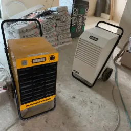 Dwa przemysłowe osuszacze powietrza, żółty Master i biały Heylo, stoją na betonowej podłodze w surowym pomieszczeniu z workami zaprawy budowlanej w tle.