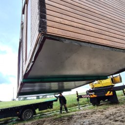 Monterdom - Transport modułowego domu z drewnianą elewacją przy użyciu dźwigu; pracownik kontroluje proces podnoszenia na błotnistym terenie.