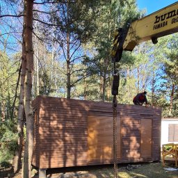 Monterdom - Montaż brązowego domu modułowego z roletami zewnętrznymi za pomocą dźwigu na zalesionej działce; pracownik w pomarańczowych rękawicach na dachu.