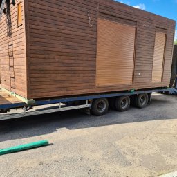 Monterdom - Transport brązowego domu modułowego z roletami zewnętrznymi na niskopodwoziowej przyczepie, widok z boku, błękitne niebo w tle.