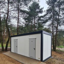 Monterdom - Biały kontener sanitarny z dwiema szarymi drzwiami, ustawiony na chodniku z kostki brukowej, otoczony drzewami iglastymi i piaszczystym podłożem.