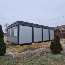 Monterdom - Gotowy, parterowy budynek z płyt warstwowych w kolorze szarym, z czarnym dachem, umieszczony na betonowych bloczkach. Widoczne rolety w oknach, trawa z resztkami śniegu i drzewa iglaste w tle.