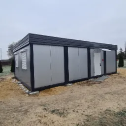 Szary pawilon modułowy z blachy falistej, z oknami z żaluzjami i białymi drzwiami wejściowymi, ustawiony na betonowych fundamentach na piaszczystym terenie w pochmurny dzień.