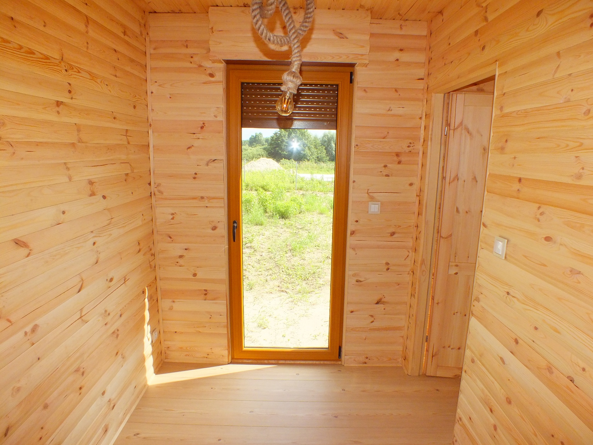 Wnętrze domu z drewnianych paneli, z widokiem na zewnątrz przez okno z roletami, ozdobione żarówką zwisającą na sznurze.