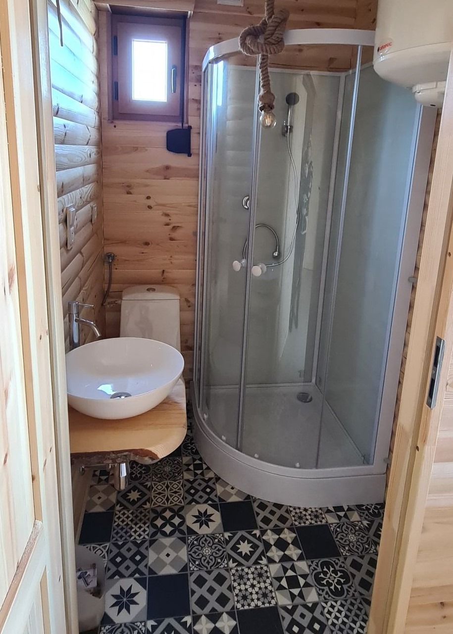 Wnętrze łazienki w domku z bali: kabina prysznicowa narożna, umywalka nablatowa na blacie z surowego drewna, toaleta, bojler i wzorzysta podłoga.