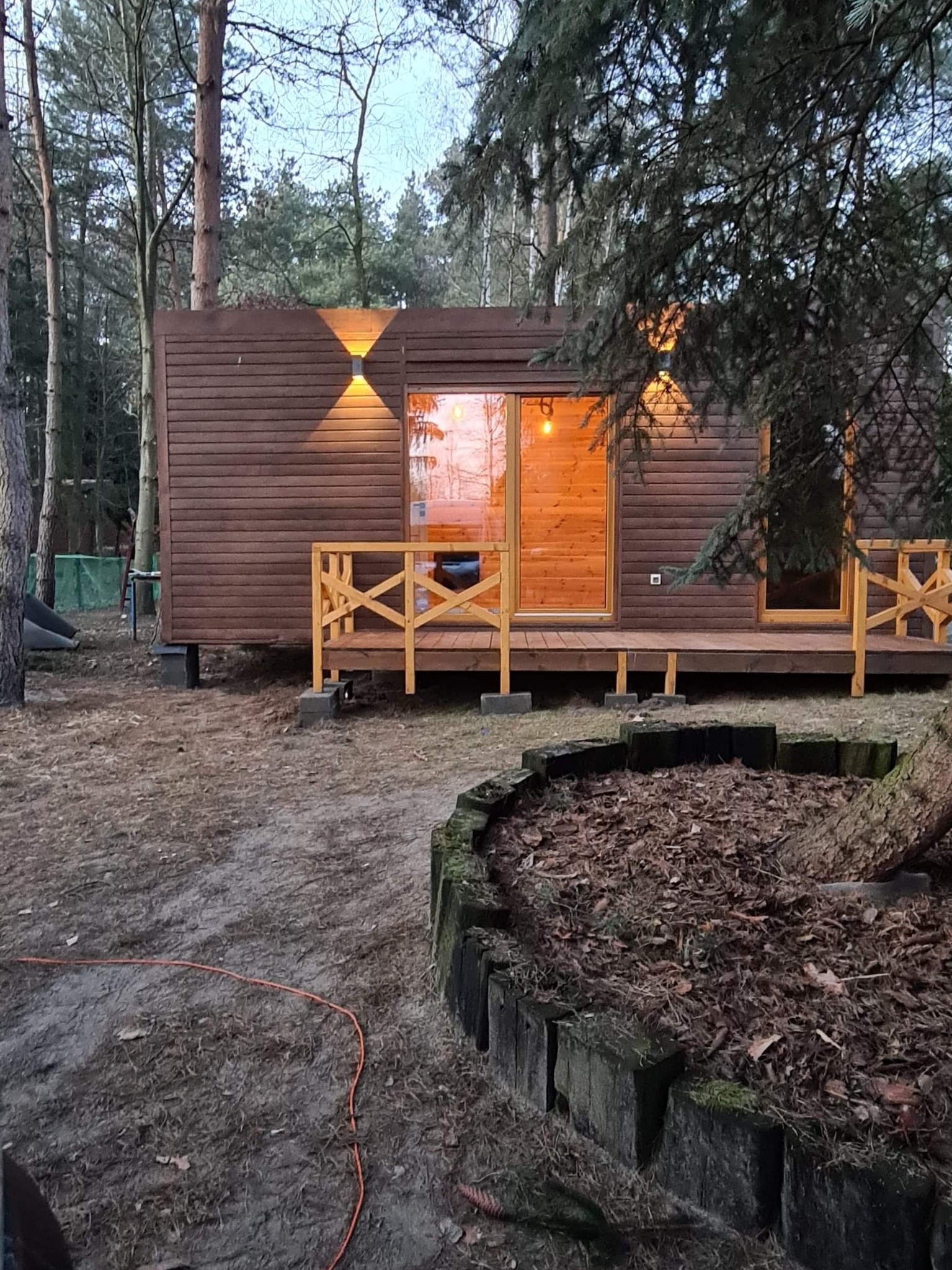 Brązowy, parterowy dom z drewnianą elewacją, oświetlony zewnętrznymi lampami i z drewnianym tarasem, umieszczony na betonowych blokach w zalesionym terenie.