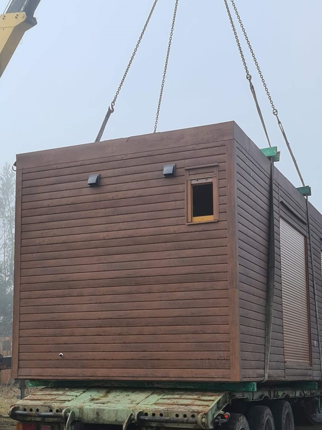 Modułowy budynek z brązową elewacją z desek, transportowany na platformie ciężarówki za pomocą dźwigu, widoczny w pochmurny dzień.