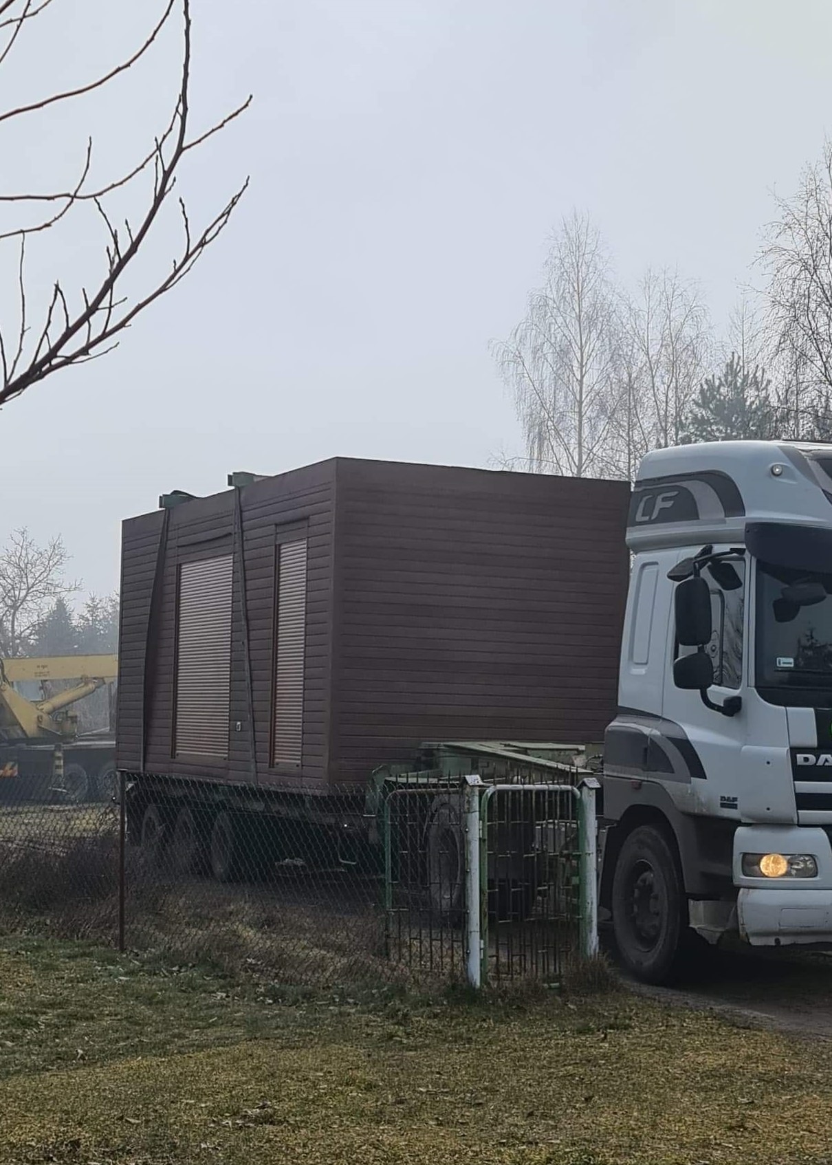 Transport modułowego domu o elewacji z brązowych paneli na naczepie ciężarówki DAF, widok zza ogrodzenia w pochmurny dzień.