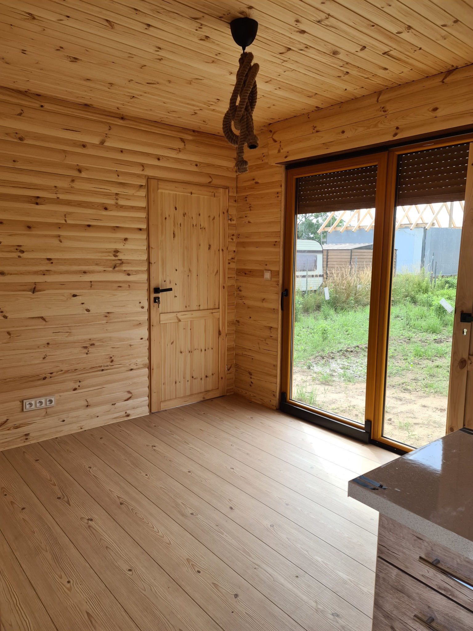 Wnętrze domu z drewnianych bali, widoczne drewniane ściany, podłoga i drzwi, okno przesuwne z roletami zewnętrznymi, oraz lampa ze sznura zwisająca z sufitu.
