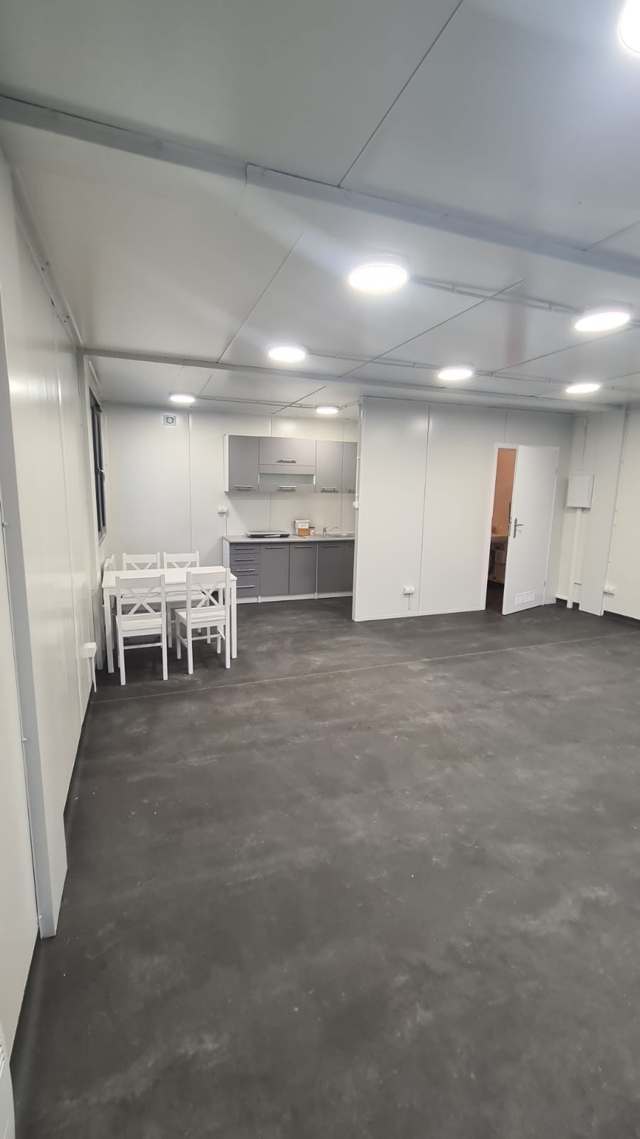 Wnętrze kontenera mieszkalnego z aneksem kuchennym w odcieniach szarości i białym stołem z krzesłami, oświetlone ledowymi panelami sufitowymi.
