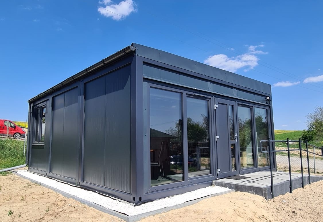 Nowoczesny, szary budynek modułowy z dużymi oknami i drzwiami, otoczony jasnym piaskiem i białym żwirem, na tle błękitnego nieba i zielonego pola.