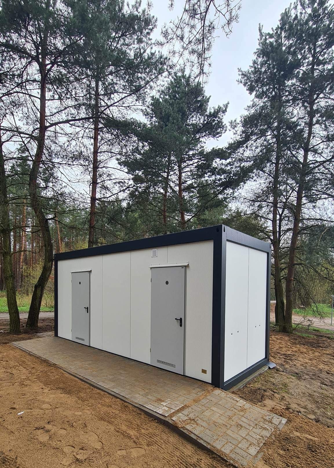 Biały kontener sanitarny z dwiema szarymi drzwiami, ustawiony na chodniku z kostki brukowej, otoczony drzewami iglastymi i piaszczystym podłożem.