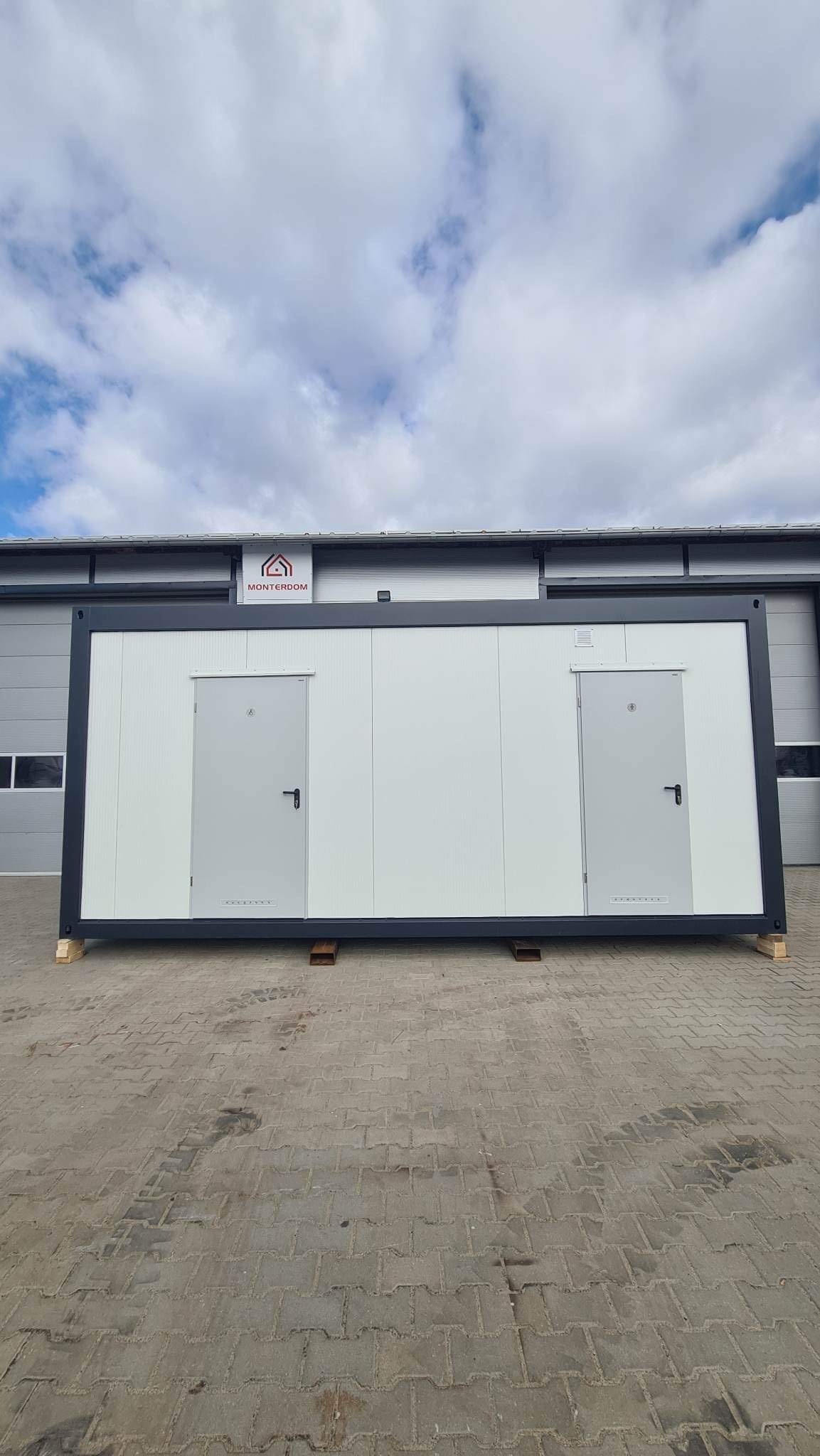 Biały pawilon modułowy z dwiema szarymi drzwiami, umieszczony na drewnianych paletach na brukowanym placu, z logo firmy na górze i budynkiem w tle pod pochmurnym niebem.
