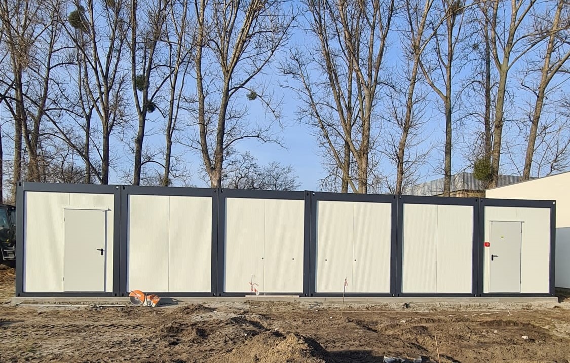 Kompleks połączonych kontenerów biurowych z drzwiami i oknami, ustawiony w Trzemesznie na placu budowy z widoczną ziemią i drzewami w tle.