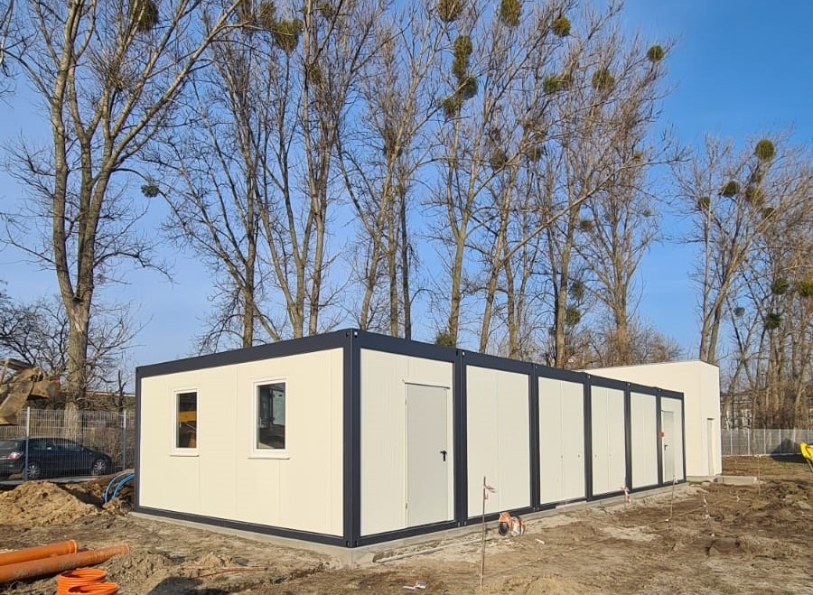 Zestaw połączonych modułów biurowych z oknami i drzwiami, teren budowy w tle z rurami i drzewami bez liści.