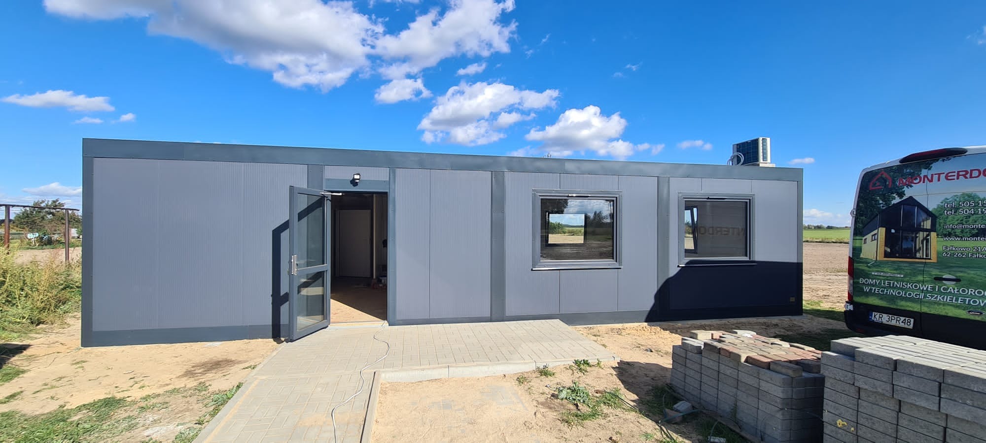 Szary budynek modułowy z otwartymi szklanymi drzwiami i dwoma oknami, widoczny klimatyzator na dachu, obok zaparkowany samochód dostawczy firmy budowlanej z reklamą domów szkieletowych.