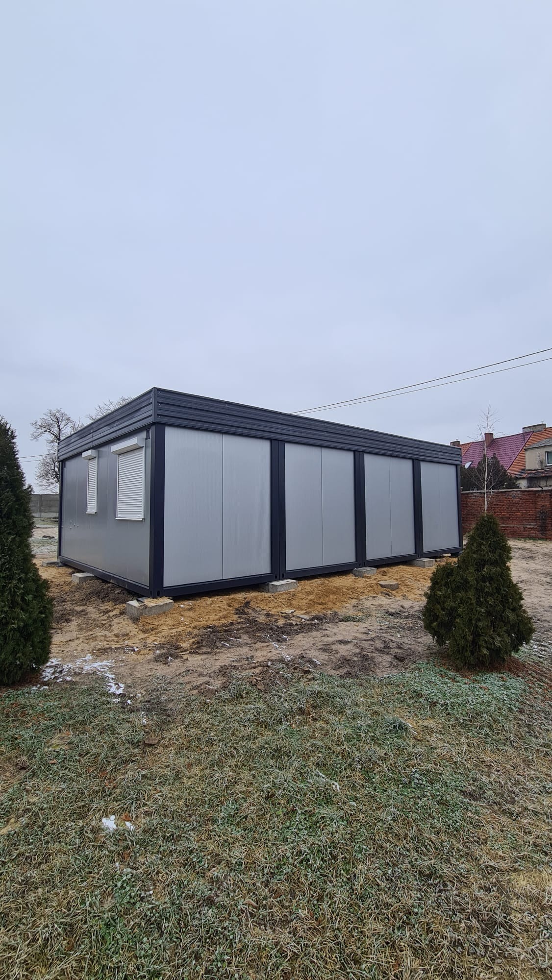 Gotowy, parterowy budynek z płyt warstwowych w kolorze szarym, z czarnym dachem, umieszczony na betonowych bloczkach. Widoczne rolety w oknach, trawa z resztkami śniegu i drzewa iglaste w tle.