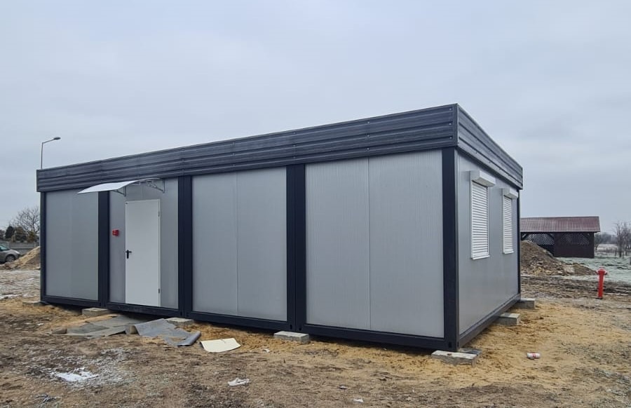 Złożony z kilku modułów szary budynek biurowy z oknem z roletą i wejściem z daszkiem, ustawiony na betonowych blokach na placu budowy.