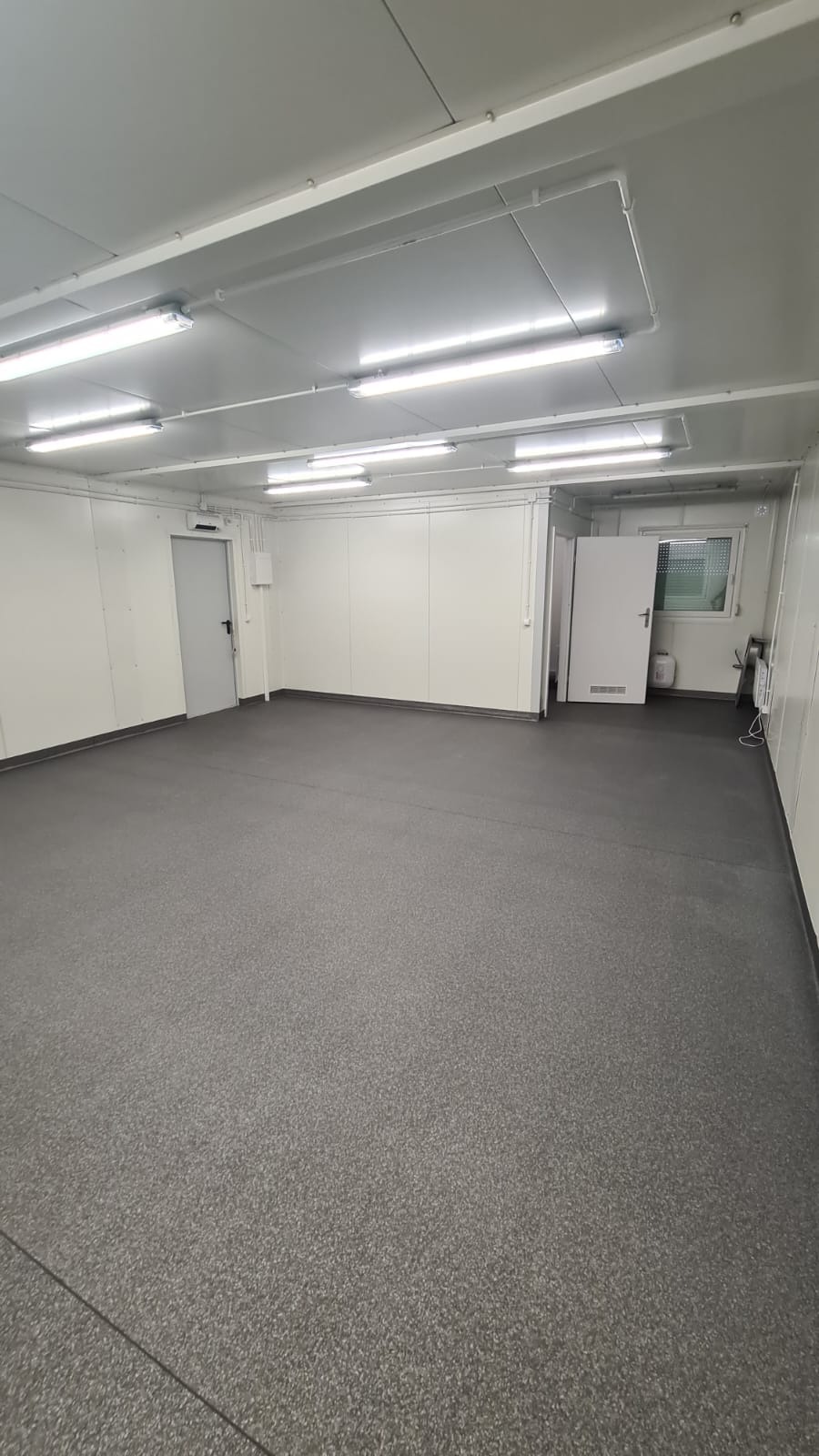Wnętrze pustego, jasnego pomieszczenia z szarą podłogą, białymi ścianami z paneli i oświetleniem jarzeniowym na suficie. Widoczne drzwi wejściowe i okno z kratą.