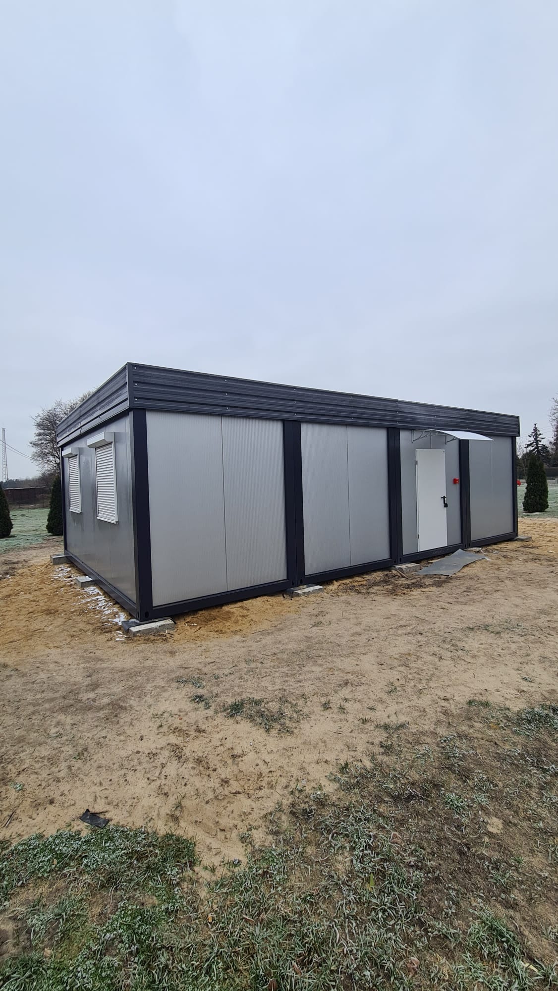 Szary pawilon modułowy z blachy falistej, z oknami z żaluzjami i białymi drzwiami wejściowymi, ustawiony na betonowych fundamentach na piaszczystym terenie w pochmurny dzień.