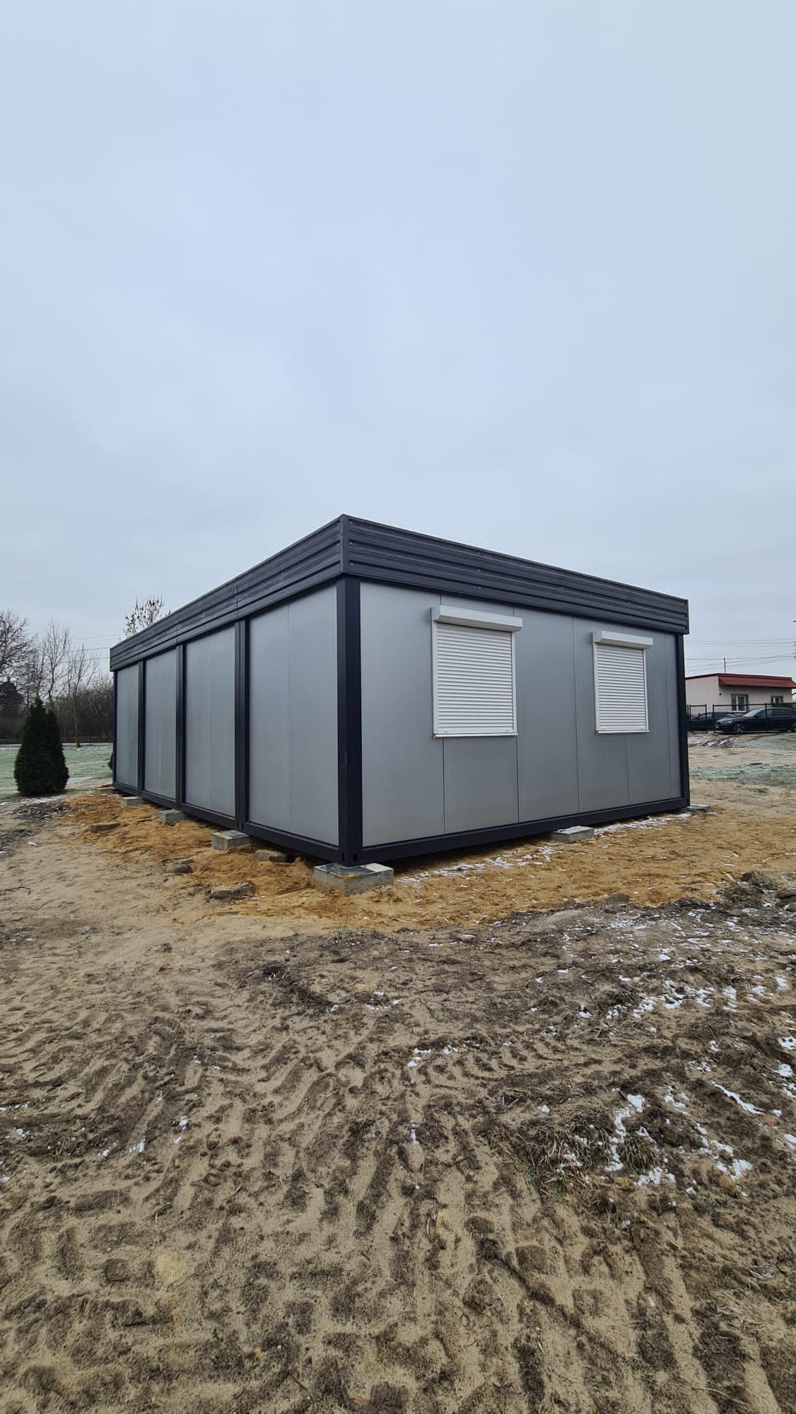 Szary budynek modułowy z roletami zewnętrznymi i czarnym obramowaniem, umieszczony na piaszczystym podłożu z widocznymi śladami opon, w tle drzewa i dom.
