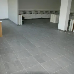 Ułożona szara podłoga z płytek w jasnym, przestronnym wnętrzu. Widoczne przygotowane elementy wyposażenia i odkurzacz przemysłowy. Minimalistyczny styl.