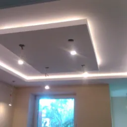 Podwieszany sufit z oświetleniem LED w kształcie kwadratów, z widocznymi kablami od lamp. Nowoczesne wykończenie wnętrza, jasne ściany i okno.