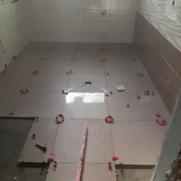 Układanie beżowych płytek podłogowych w łazience z użyciem systemu poziomowania, widoczny n&oacute;ż do cięcia płytek, częściowo ułożona podłoga i ściany wyłożone płytkami z wzorem fal.