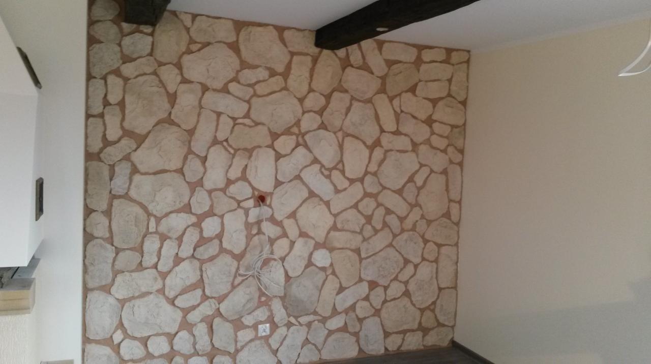 Ściana wykończona dekoracyjnym tynkiem imitującym kamień, z widocznymi elementami instalacji elektrycznej i fragmentami drewnianych belek stropowych.