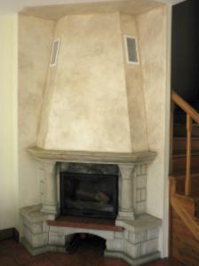 Ozdobny kominek w rogu pomieszczenia, wykończony w jasnych odcieniach beżu i szarości, z widoczną strukturą na powierzchni. Kominek obudowany kamiennymi elementami, z drewnianymi schodami w tle.