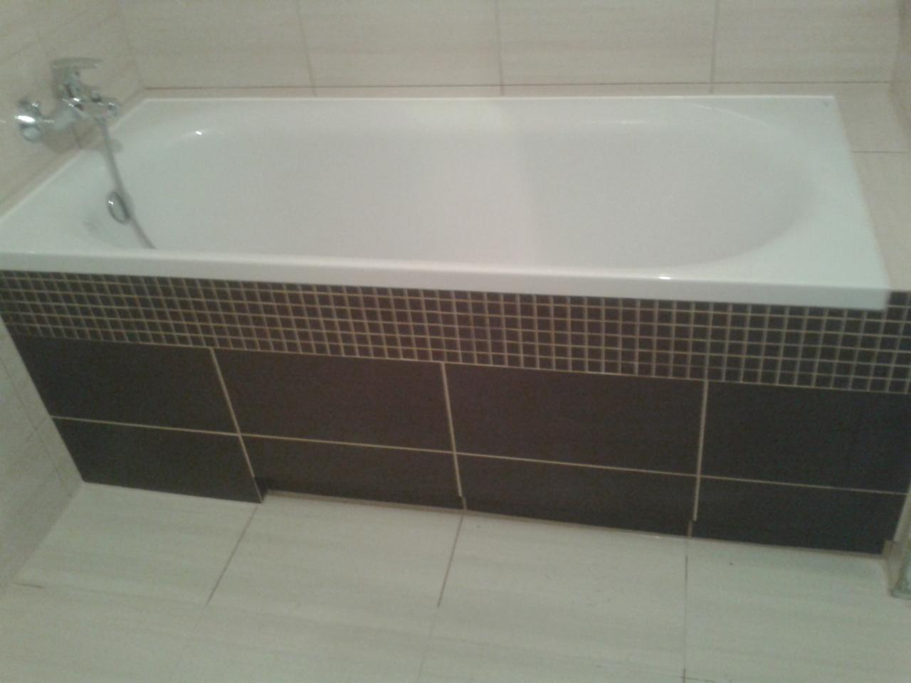 Biała wanna obudowana ciemnymi płytkami z dekoracyjnym pasem mozaiki, zamontowana bateria, jasna podłoga i ściany wyłożone płytkami.