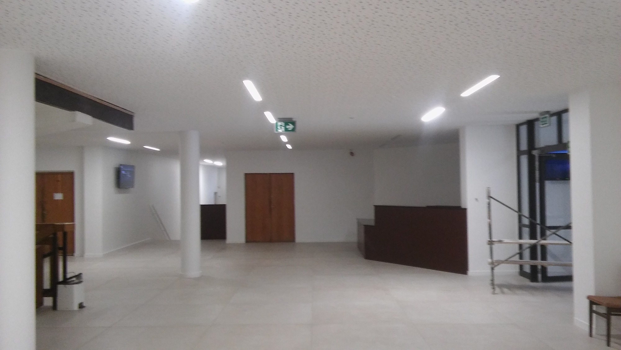 Jasne, przestronne wnętrze biurowe z białymi ścianami, kolumnami i podwieszanym sufitem z oświetleniem LED, widoczne drzwi i fragmenty wyposażenia.